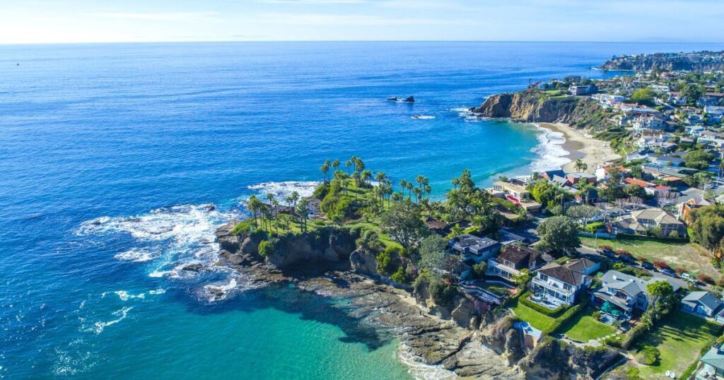 An aerial view of Laguna Beach, showcasing its stunning coastal landscape with lush palm trees, rocky outcrops, and azure waters. The sandy beach curves gently along the neighborhood, with houses perched on the cliffs offering panoramic views of the Pacific Ocean. This picturesque scene highlights why Laguna Beach is celebrated as one of the best day trips from LA, offering a perfect blend of natural beauty and luxury living.