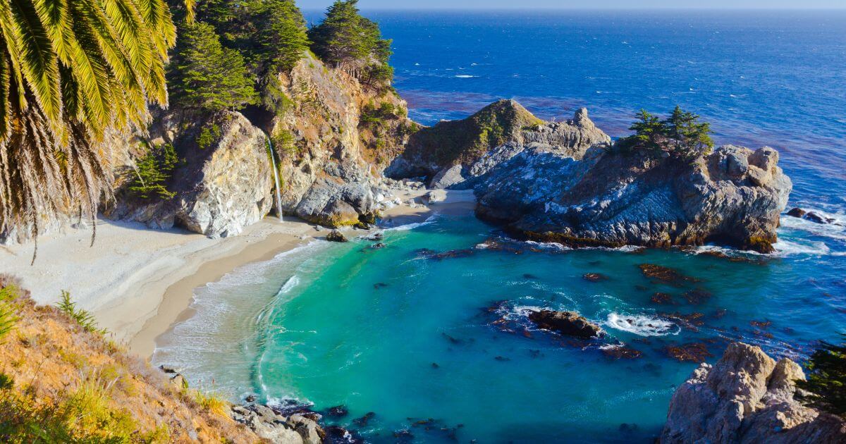 Breathtaking view of McWay Falls, where a cascading waterfall drops directly onto a pristine sandy beach, surrounded by rugged cliffs and rock formations. Turquoise waters gently lap the shoreline, and tall trees frame the scene on the left.