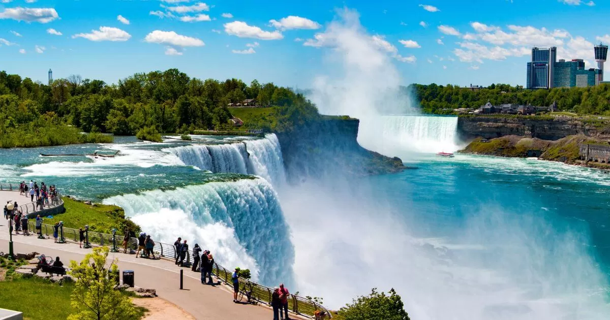 Breathtaking view of Niagara Falls on a clear day, with cascading water, a boat navigating the misty waters below, and visitors observing from a nearby viewing platform, with city skyline in the distance.