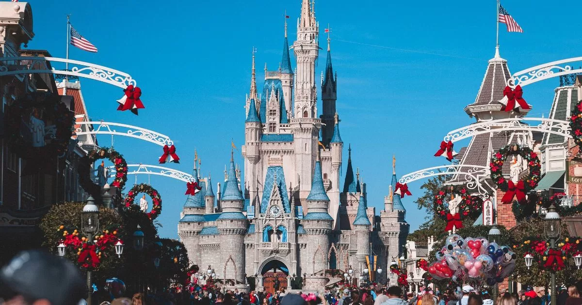 Festive scene at Walt Disney World with Cinderella's Castle standing majestically in the background, adorned with vibrant holiday decorations. Red bows and garlands line the overhead arches and buildings, while visitors bustle about, some carrying colorful Disney-themed balloons.