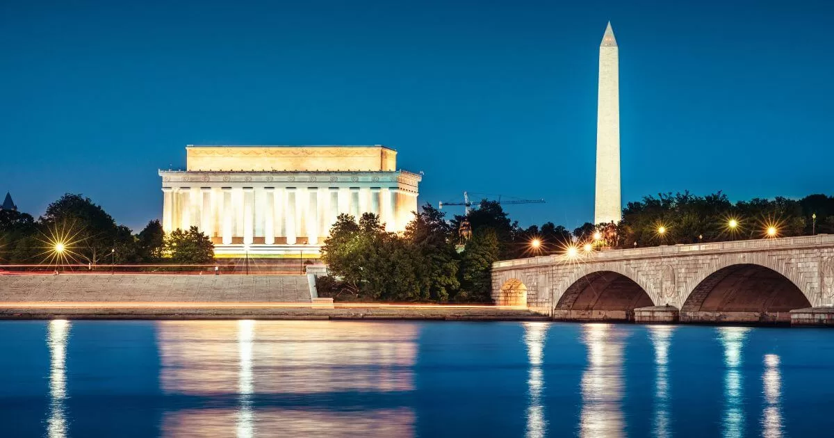 Evening view of Washington DC, one of the best places to visit in the USA, with the illuminated Lincoln Memorial on the left and the towering Washington Monument on the right. Both landmarks reflect softly on the tranquil waters of the Potomac River, which is spanned by a stone arched bridge adorned with glowing lampposts.