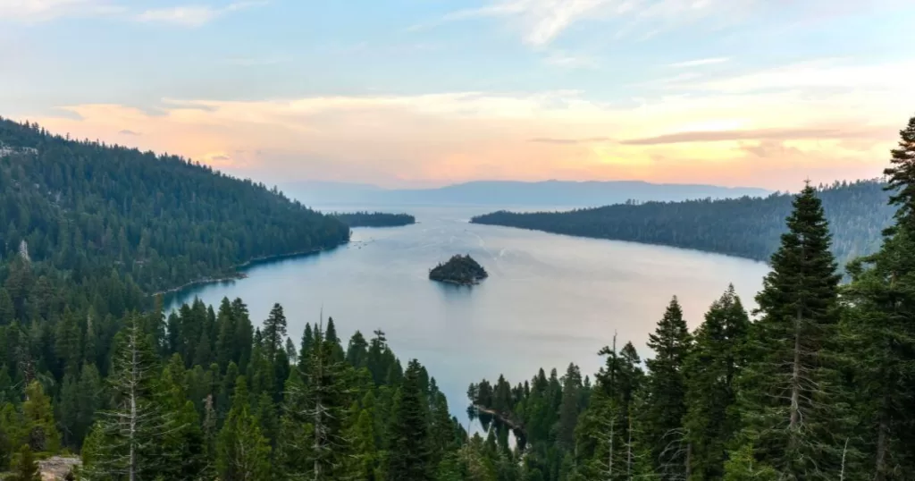 The serene Emerald Bay State Park at dusk, showcasing one of the best camping sites in California. The bay is captured in a tranquil state with smooth waters reflecting the pastel hues of the sunset. Fannette Island, the only island in Lake Tahoe, is centrally located in the bay, adding to the picturesque scenery. Surrounding the bay, lush green forests of tall pine trees create a natural amphitheater, perfect for nature enthusiasts and campers seeking a peaceful retreat amidst stunning landscapes. The fading light casts a soft glow over the scene, suggesting the calming end to a day of outdoor adventure.