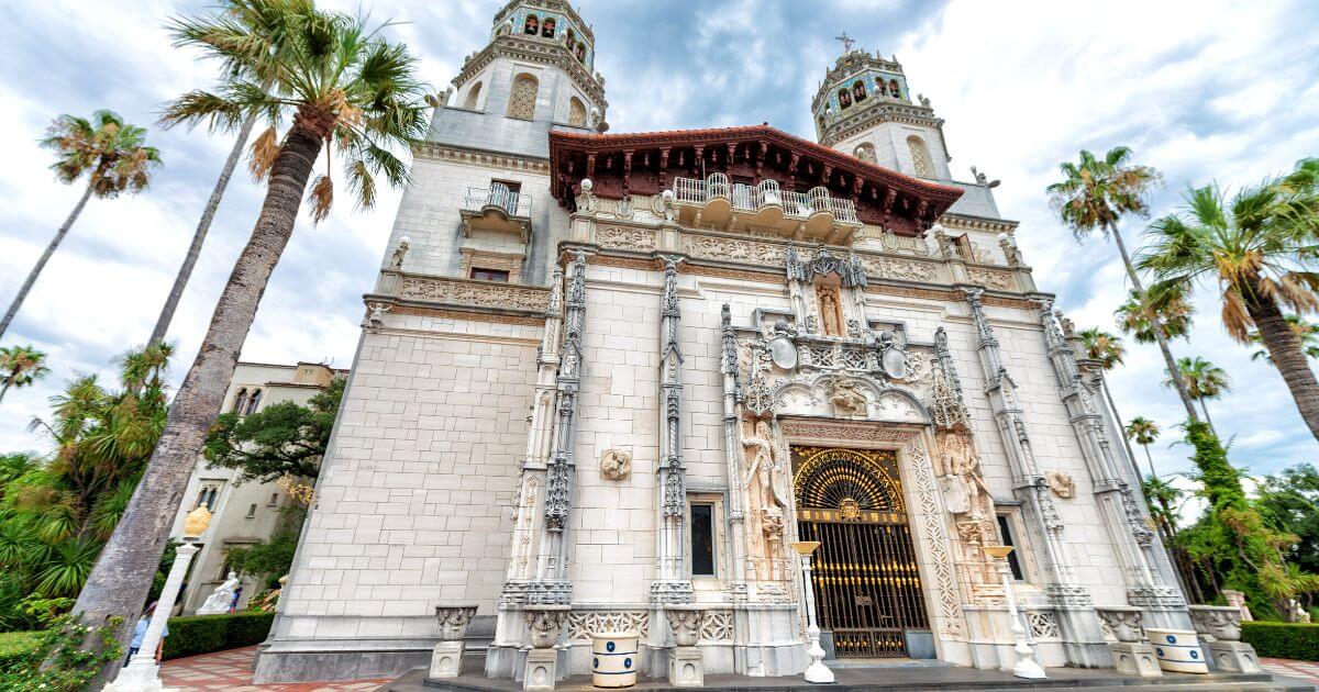 Hearst Castle facade featuring intricate architectural details, tall palm trees, and ornate entrance gates against a cloudy sky backdrop.