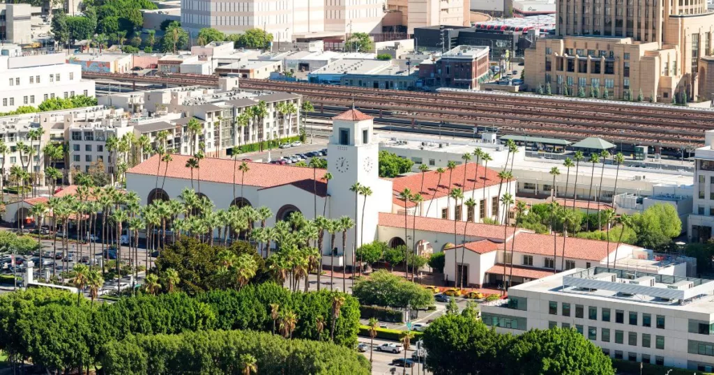 Los Angeles Union Station viewed from above, with its iconic white bell tower, surrounded by palm trees and set against the city's bustling railway tracks and urban backdrop.