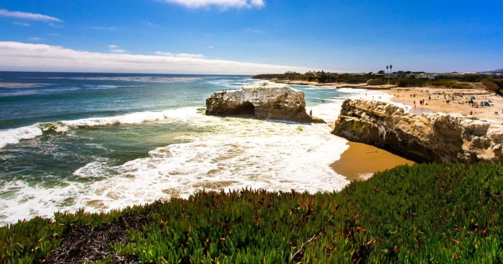 Vibrant beach scene at Santa Cruz, California, with robust waves crashing against a rocky outcrop and beachgoers enjoying the sun-kissed shore.