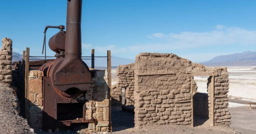 Ruins of the Harmony Borax Works in Death Valley, with a large, rusted boiler and remnants of adobe walls against a backdrop of distant mountains and salt flats.