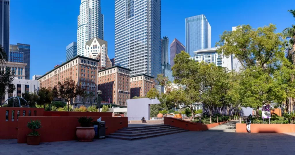 Pershing Square in downtown Los Angeles, a public space with walkways, seating areas, and greenery set against a backdrop of towering skyscrapers and clear blue skies.