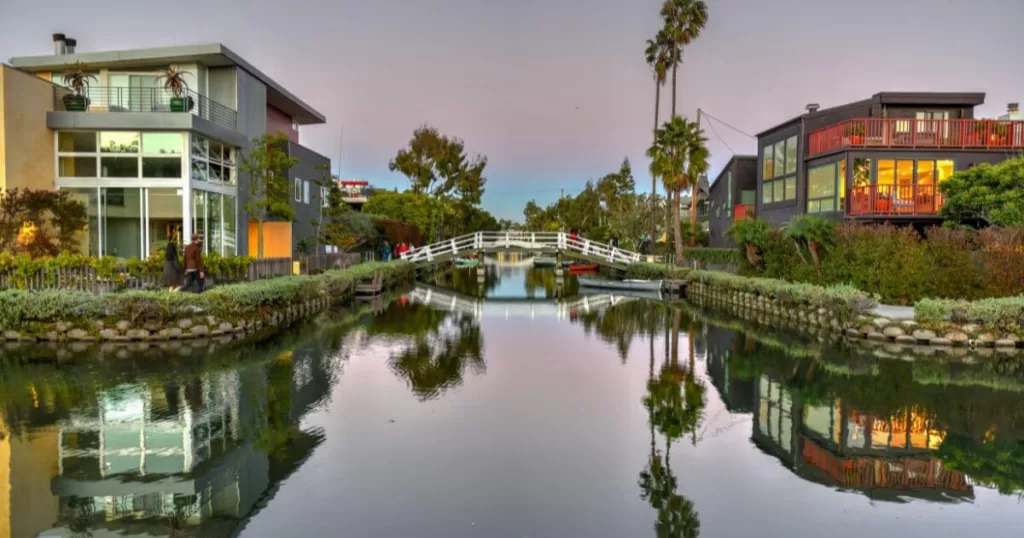 Twilight over the Venice Canals in Los Angeles, reflecting modern homes and palm trees on calm waters, with a pedestrian bridge connecting the tranquil pathways.