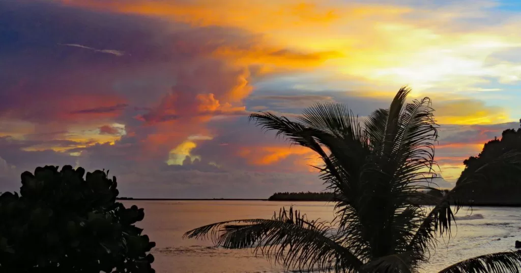 A serene sunset in American Samoa, one of the best national parks in the United States, with radiant clouds in shades of orange and purple above the tranquil sea, silhouetted palm trees, and lush tropical vegetation.