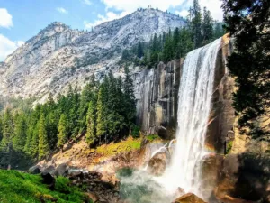 Majestic Vernal Falls in full flow, one of the most beautiful waterfalls in California, with mist rising above rocky terrain surrounded by lush greenery and towering cliffs.