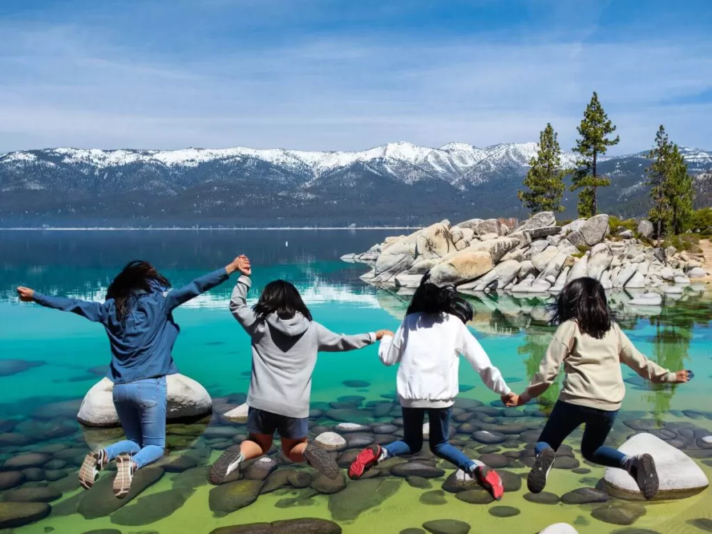 Four children holding hands and jumping over rocks at Lake Tahoe's clear shore with snow-capped mountains in the background, enjoying things to do in California with kids.