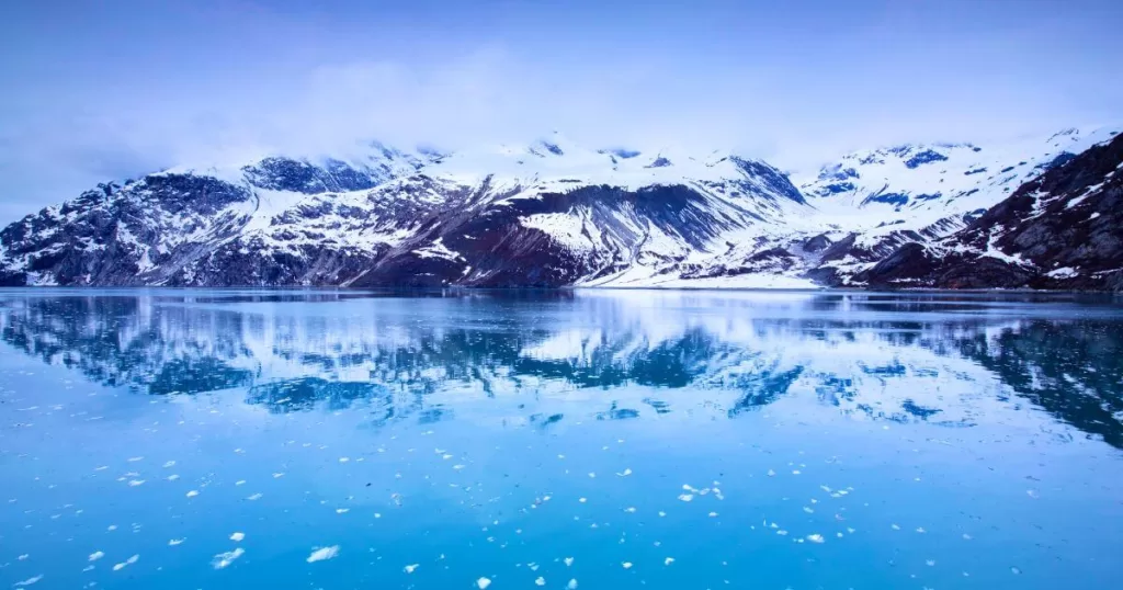 Glacier Bay National Park, one of the best national parks in the United States, presents a tranquil scene with a mirror-like reflection of snow-capped mountains and scattered ice on the calm blue waters, under a soft overcast sky.