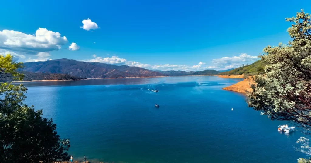 A panoramic view of Lake Shasta, with its deep blue waters surrounded by mountains and lush greenery, with boats leisurely cruising and people enjoying the shoreline.