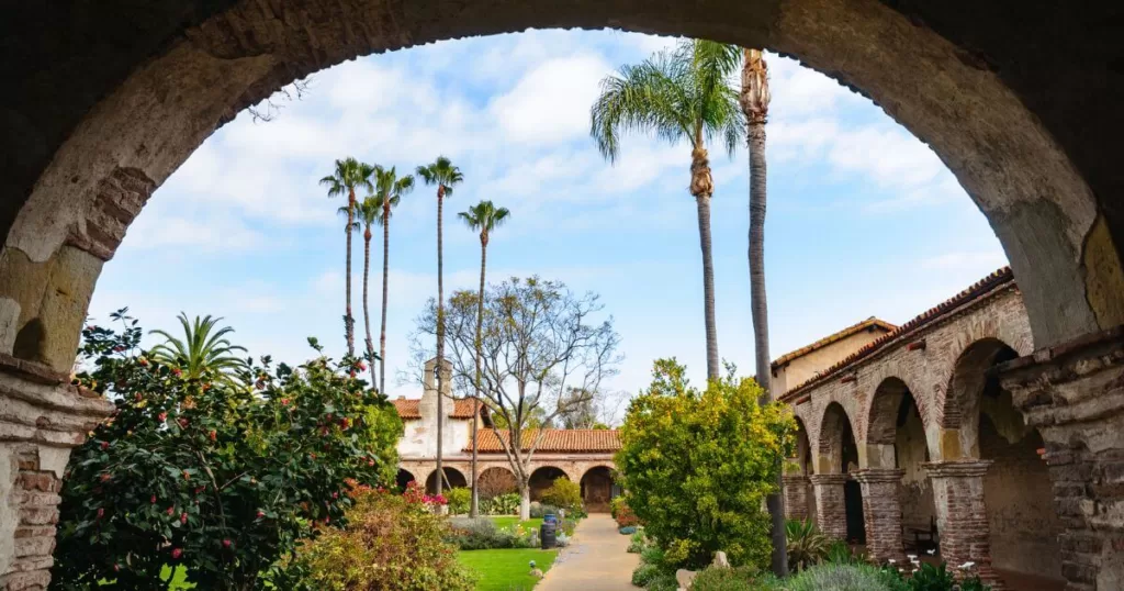 View through an arched passageway at Mission San Juan Capistrano, showcasing the historic mission's aged brickwork and elegant arches. Lush gardens filled with flowering plants, tall palm trees, and greenery frame the courtyard, under the open sky, highlighting the serene and timeless beauty of this well-preserved Spanish mission in Southern California.