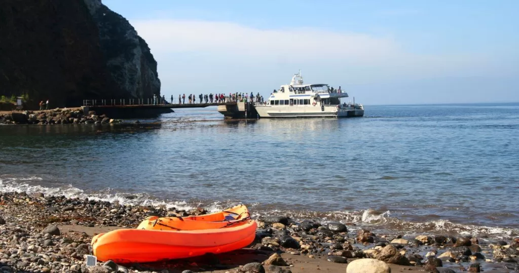 A serene view of Santa Cruz Island's landing area with a ferry boat docked at the pier and a group of visitors ready to explore the island. In the foreground, an orange kayak rests on the pebbly beach, inviting adventure on the calm sea waters, with the rugged cliff of the island providing a majestic backdrop under a clear blue sky.