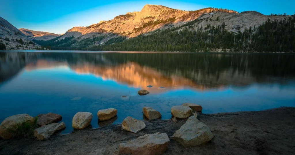 Twilight descends on Tenaya Lake, one of the best things to do in Yosemite National Park, with the tranquil waters mirroring the soft sky and the alpenglow on the surrounding granite peaks. Boulders line the serene lakeshore, enhancing the peaceful and reflective ambiance of the scene.