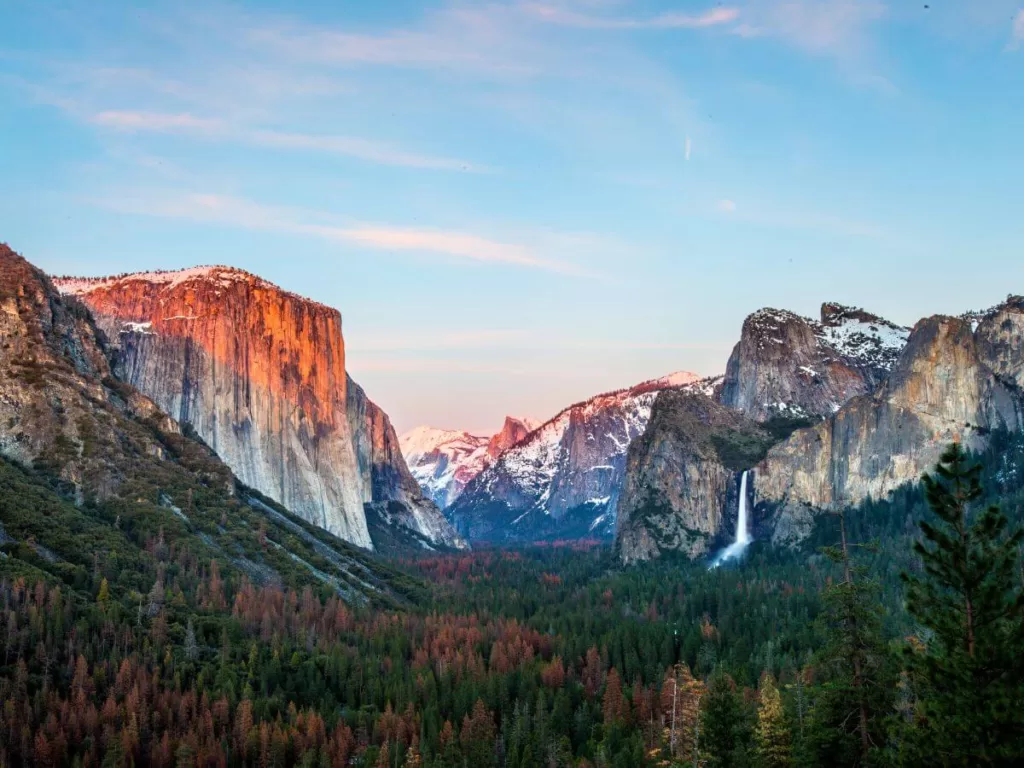 osemite's iconic Tunnel View at dusk, showcasing the breathtaking grandeur of one of the best national parks in the United States. El Capitan stands majestically on the left with its vertical granite face catching the last rays of the sun, while Bridalveil Fall cascades down the cliff on the right. Snow-dusted peaks in the background and a lush valley with pine trees complete this stunning natural vista.