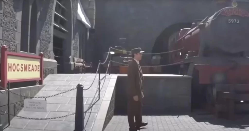 A conductor stands by the Hogsmeade station sign with the iconic red engine in the background at Universal Studios Hollywood, a magical experience for visitors.