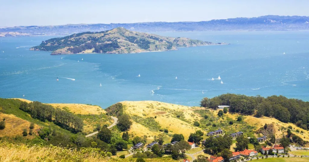 A panoramic view of Angel Island State Park with its rolling hills and patches of woodland, overlooking the sparkling blue waters of the San Francisco Bay dotted with sailboats.