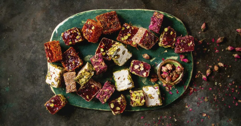 An assortment of Aplets and Cotlets Candy Kitchen's Turkish delights, arrayed on a green plate with sprinkled pistachios and rose petals, showcasing the sweet treats' vibrant colors and textures.