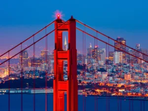 The Golden Gate Bridge at twilight with its 'International Orange' color standing out against the San Francisco skyline, illuminated by thousands of lights. A clear view from the bridge showcases the dense cityscape and iconic structures, highlighting things to do in San Francisco.