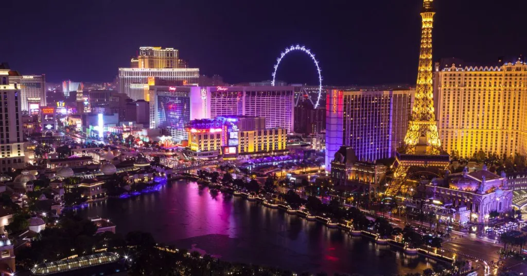 Nighttime view of the Las Vegas Strip, illuminated by the vibrant neon lights of casinos, the iconic replica Eiffel Tower, and the High Roller observation wheel, reflecting the city's bustling nightlife.