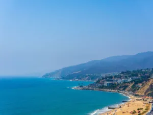 A panoramic view of Malibu beach highlighting things to do in Malibu, with sunbathers on the sand, azure waters, and cars parked near the Pacific Coast Highway, framed by green hills and houses, under a bright blue sky.