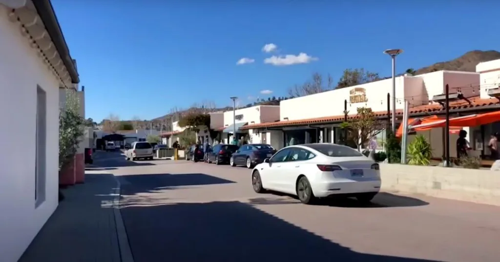 
A sunny day at Malibu Country Mart with visitors strolling and cars parked along the boutique shops, against a backdrop of green hills under a clear blue sky.