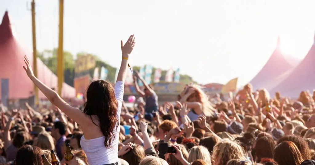 Euphoric crowd at a music festival, with a sea of fans raising their hands towards the sunny sky. The festival atmosphere is alive with energy, punctuated by the iconic pointed tops of festival tents in the background, capturing the essence of live music events and the shared joy they bring.