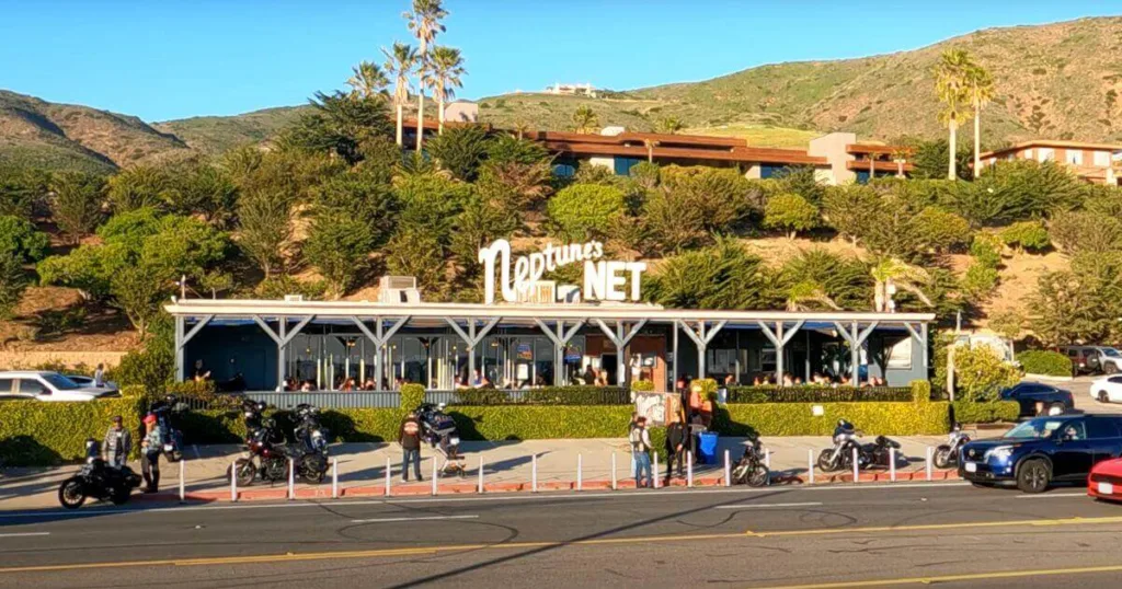 The famous Neptune's Net seafood restaurant, a popular stop for things to do in Malibu, with a lively crowd and motorcycles parked in front, set against the backdrop of Malibu's rolling hills.