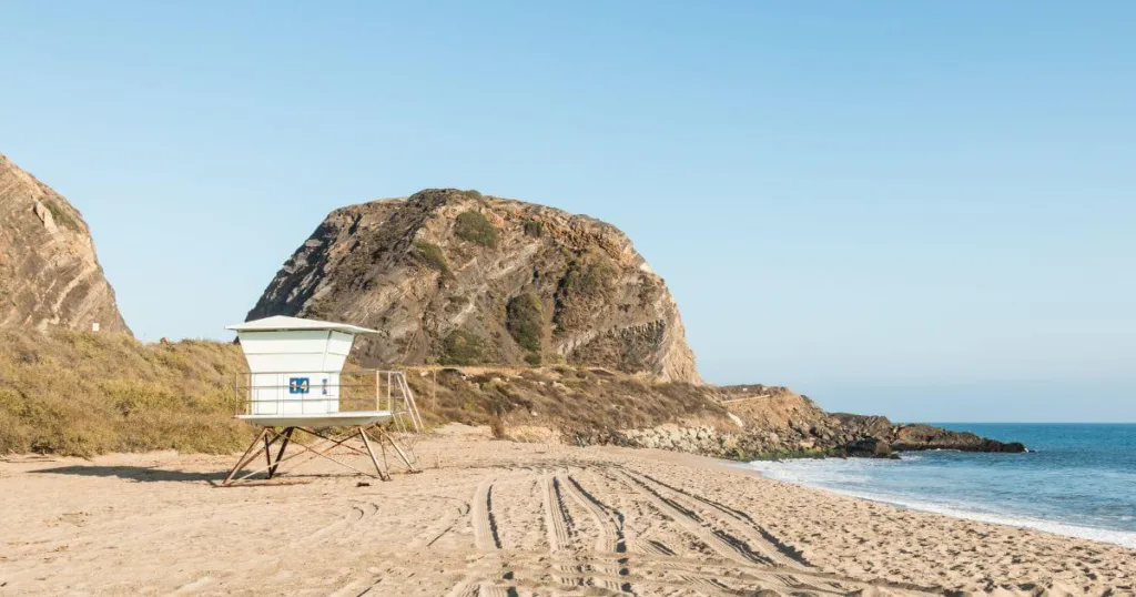 A tranquil scene at Point Mugu State Park with a lifeguard tower on a sandy beach, tire tracks marking the sand, and a large rugged cliff in the background, under a clear blue sky.