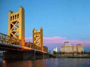 A serene evening view of Sacramento showcasing the Tower Bridge, a golden-lit vertical lift bridge, spanning across the Sacramento River. The deepening blue sky is tinted with hues of pink and orange at sunset, creating a picturesque backdrop for one of the many things to do in Sacramento. In the distance, the city skyline presents a harmonious blend of modern and historic architecture, inviting exploration and discovery.