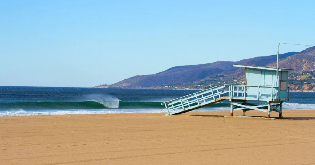 A serene day at Zuma Beach with a clear sky, showcasing a solitary lifeguard tower on the sandy shore, gentle surf waves, and the rolling hills of Malibu in the background.