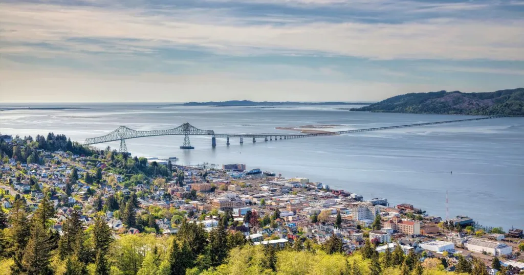 Overlooking Astoria, Oregon, this image captures the charming coastal town with its dense clusters of homes and buildings, set against the backdrop of the expansive Astoria-Megler Bridge stretching across the Columbia River, with rolling hills and the vast water body framing the scenic landscape.