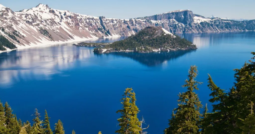 A breathtaking view of Crater Lake's deep blue waters with Wizard Island in the foreground, surrounded by steep cliffs under a clear blue sky, showcasing one of the most majestic places to visit in Oregon.