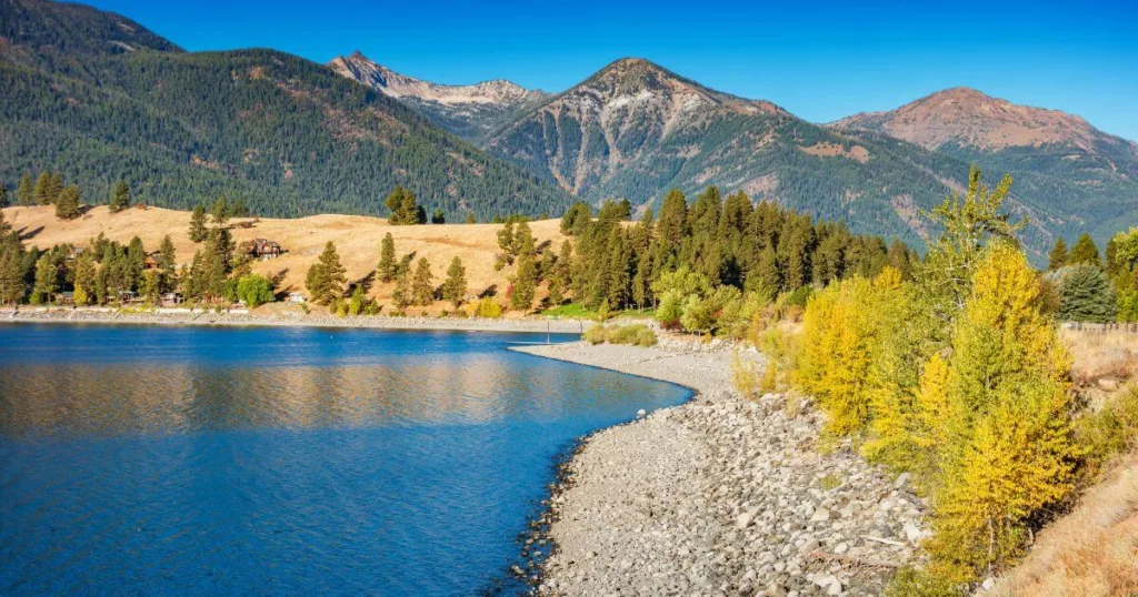 A vibrant autumn view of Wallowa Lake with clear blue waters, surrounded by golden and evergreen trees against a backdrop of the majestic Wallowa Mountains.